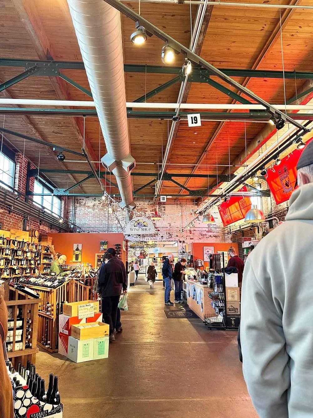 Vineyard wine shop interior with shoppers, industrial ceiling, brick walls, wine displays, and vibrant lighting.