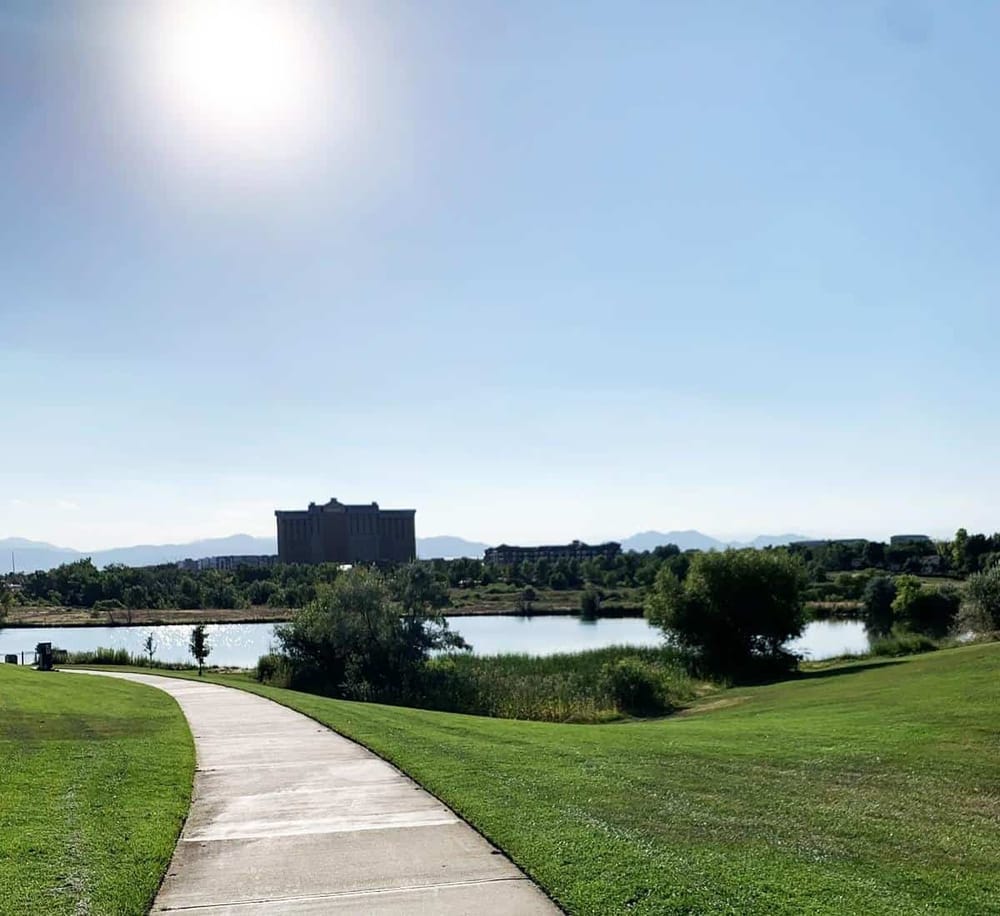 Quiet lakeside park with walking path, greenery, and city skyline in the background.