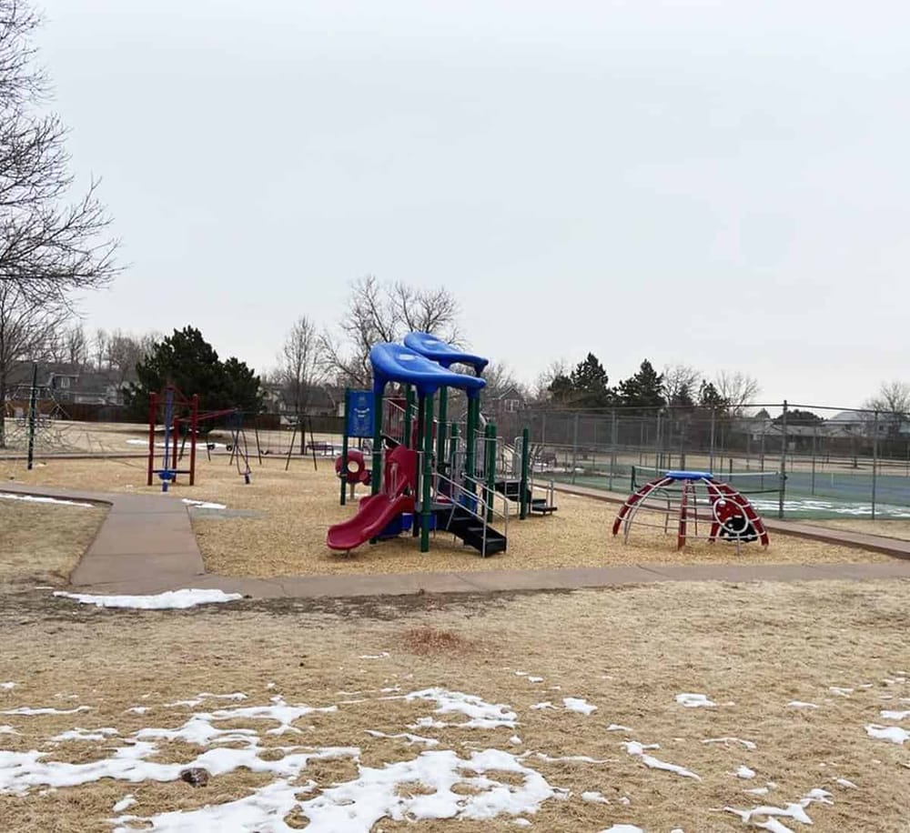 Colorful children's playground equipment at outdoor park with tennis courts and trees.