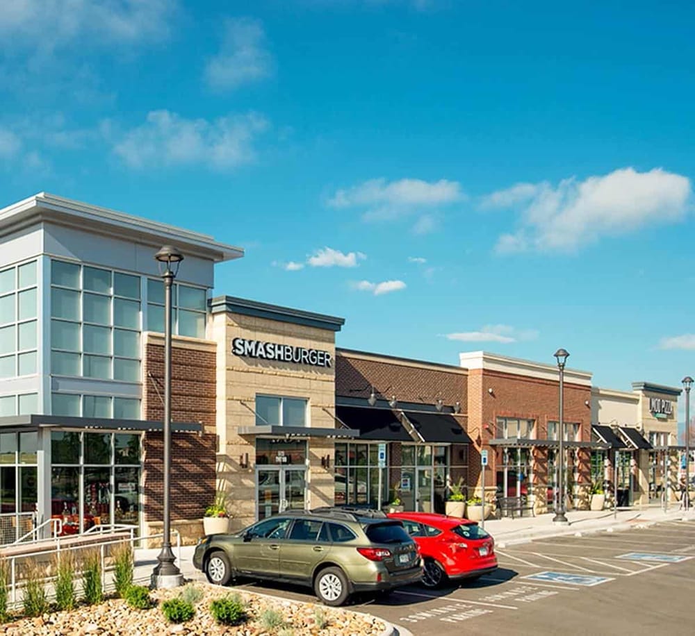 Modern shopping center with Smashburger restaurant and retail stores, outdoor parking, bright blue sky.