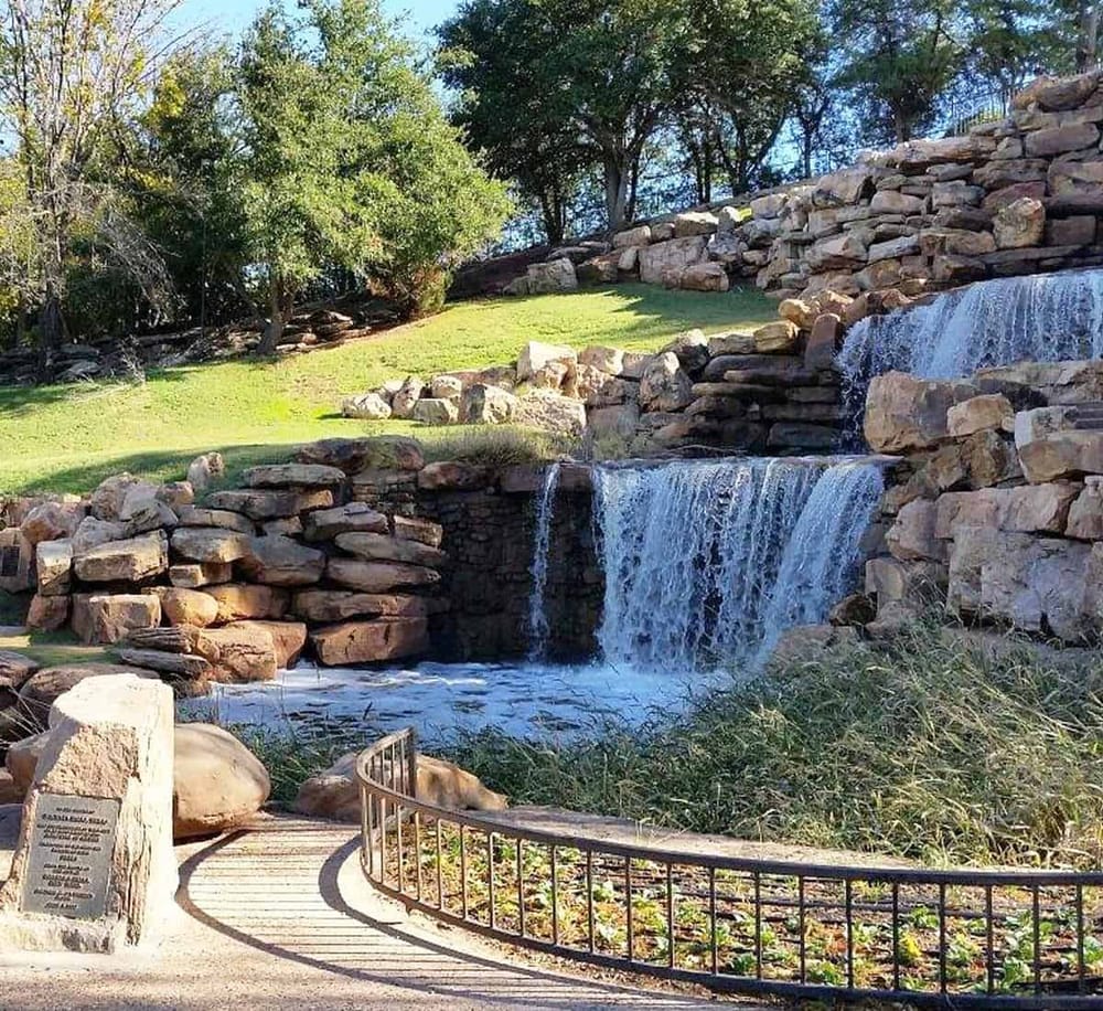 Serene waterfall on landscaped rocks at a scenic outdoor park with trees and gravel pathway.