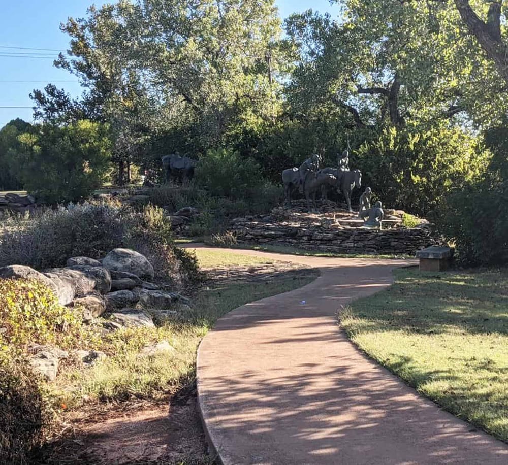 Bronze statues of cowboys and cattle on a landscaped park trail with trees and rocks.