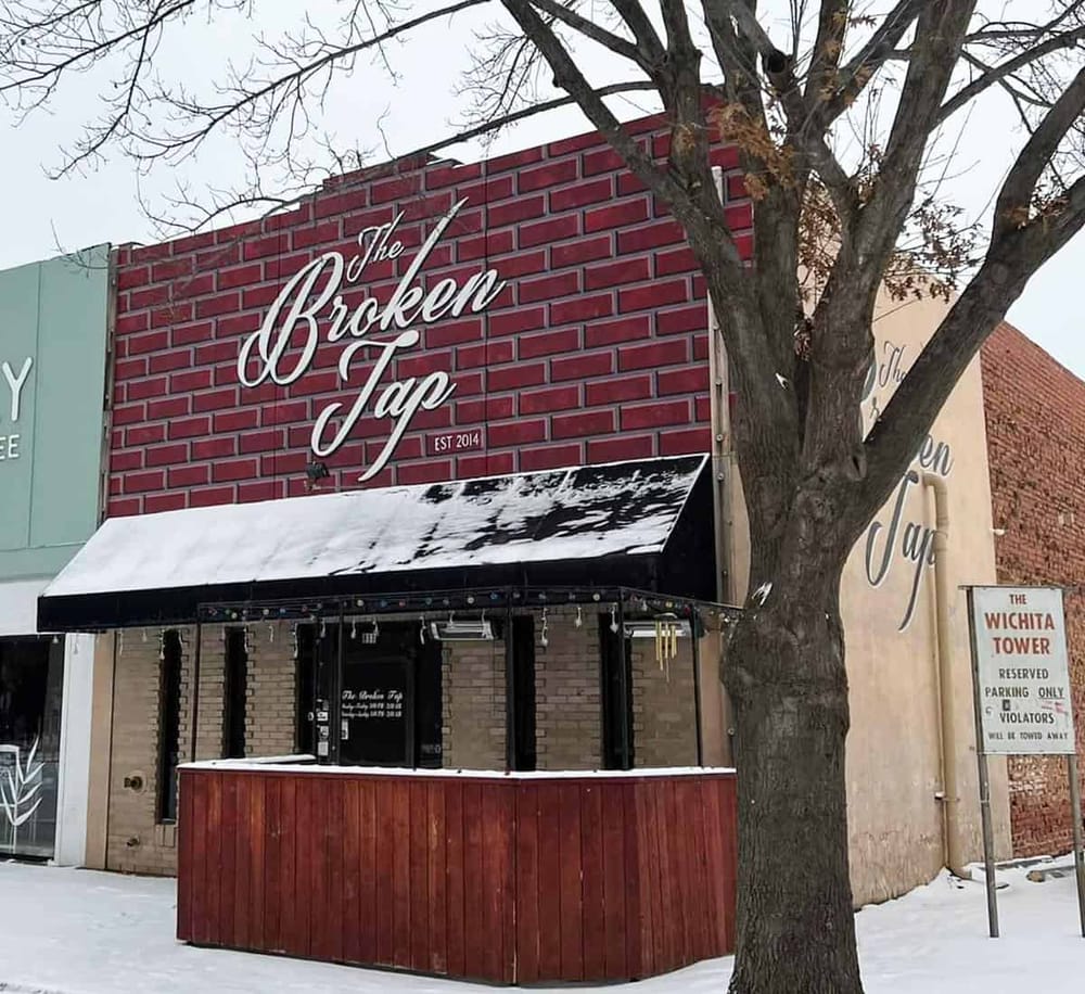 Cozy bar storefront with brick wall sign reading "The Broken Tap" in winter, snow-covered outdoor seating area, urban street scene.