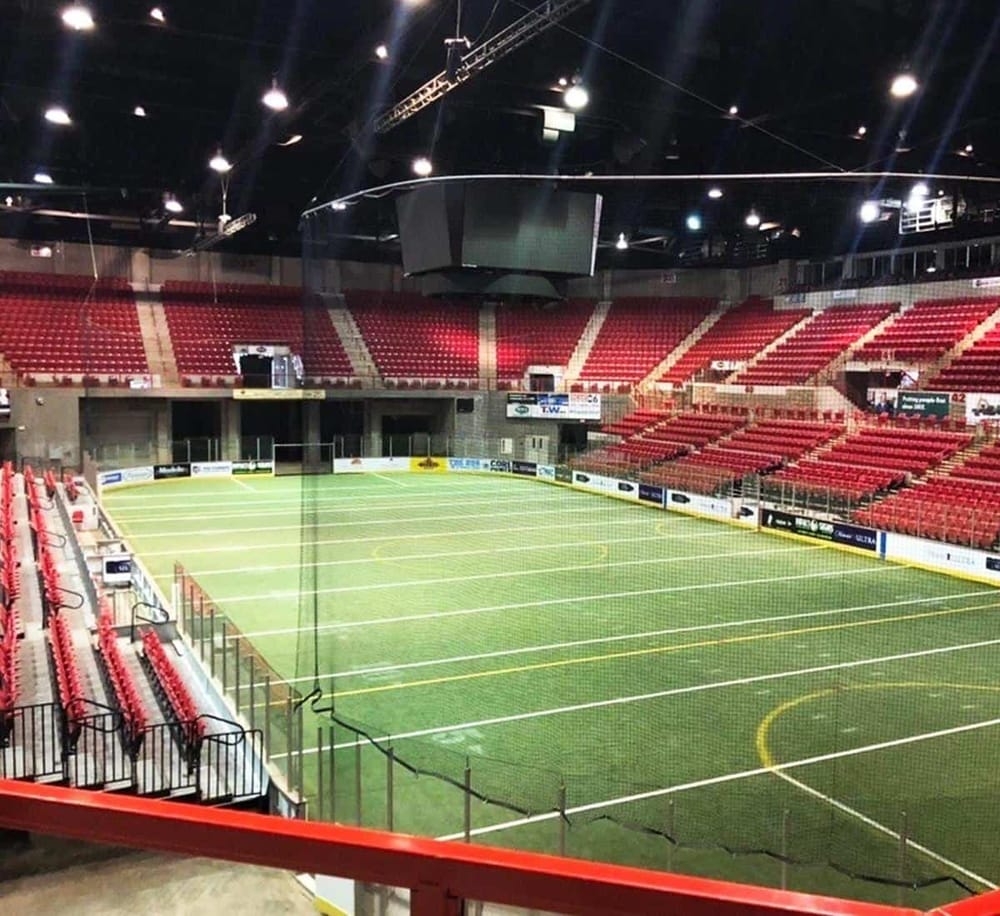 Indoor sports arena with a green turf field, red seating, and large screens for sports events.