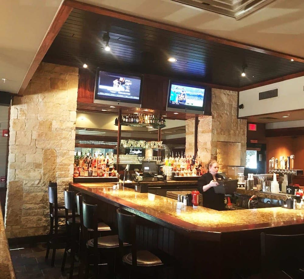 Bright hotel bar with TV screens, alcohol bottles, and a bartender in a welcoming setting.