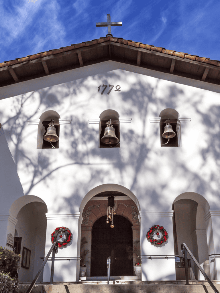 Bell tower of historic church, built in 1772, with white walls, arched entrance, and decorative wreaths, under a bright blue sky.