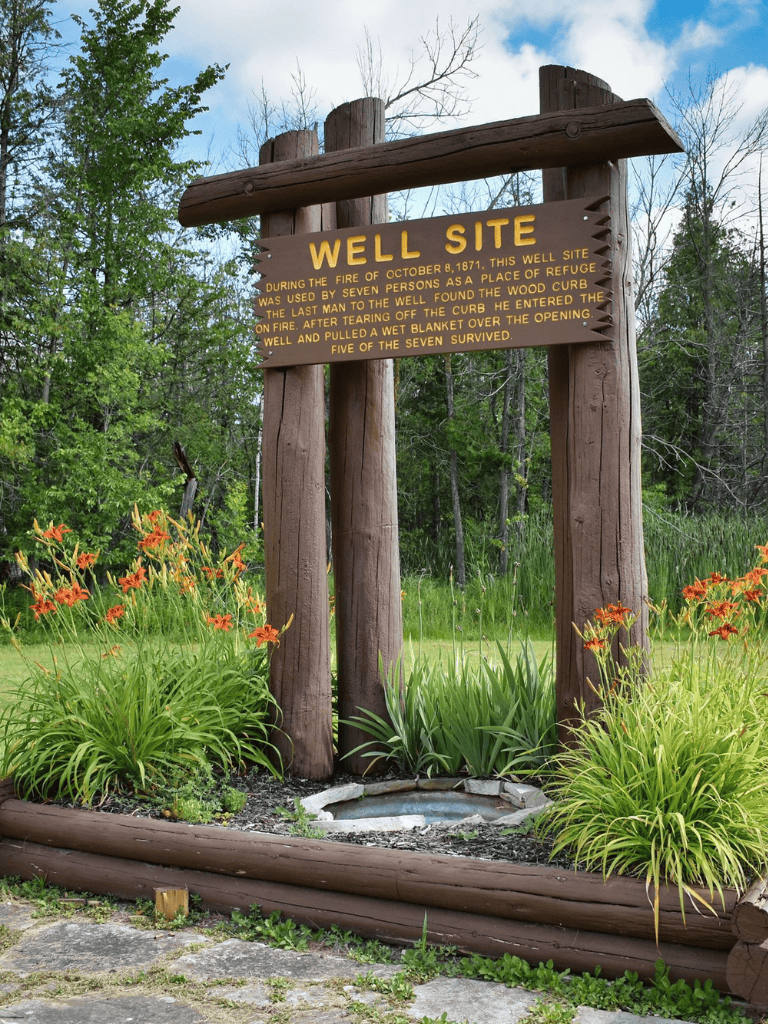 A0: Historic well site marker with wooden sign, surrounded by lush greenery and orange flowers.
