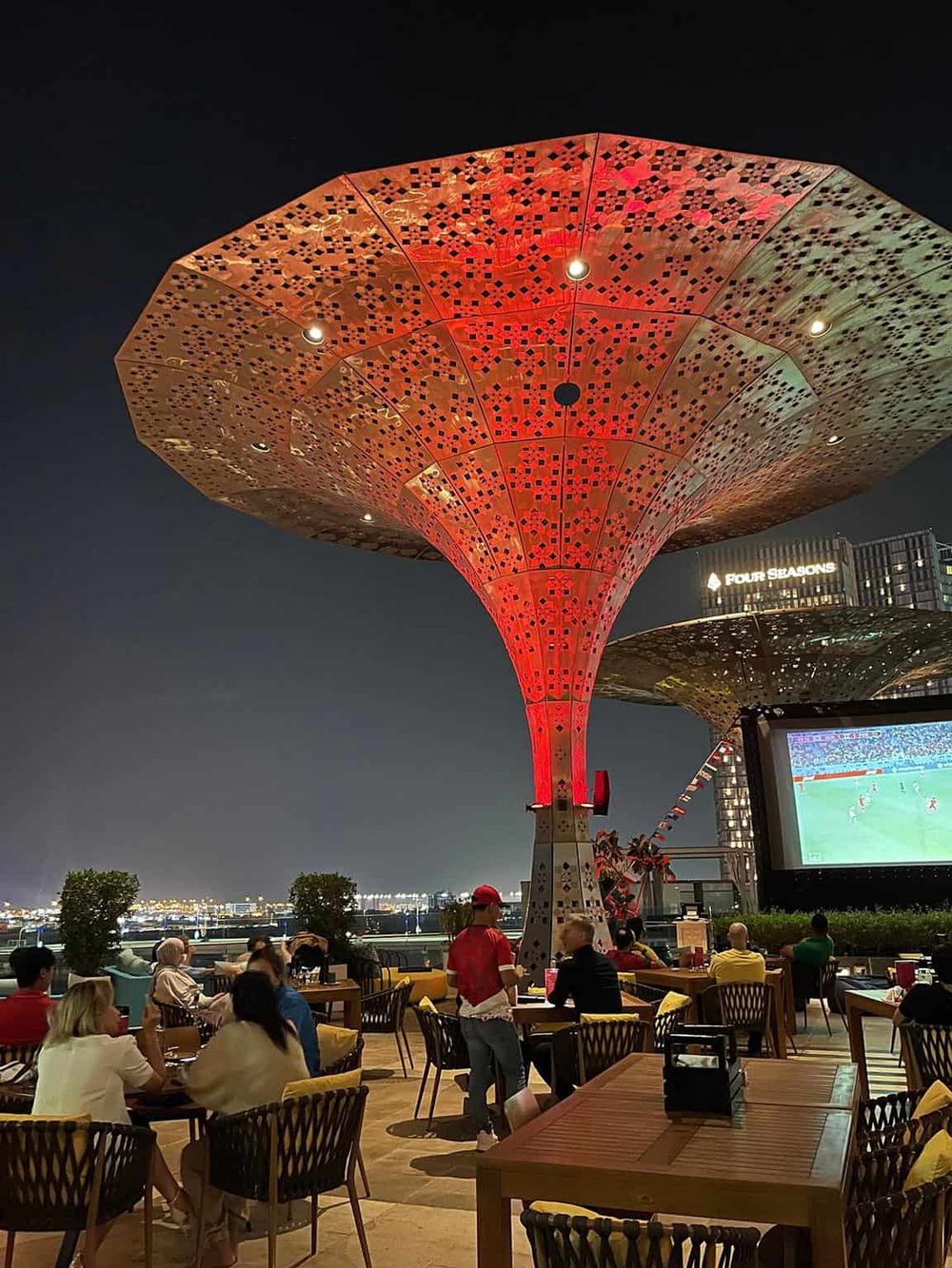 Vibrant outdoor rooftop bar with large illuminated art installation and city skyline views.