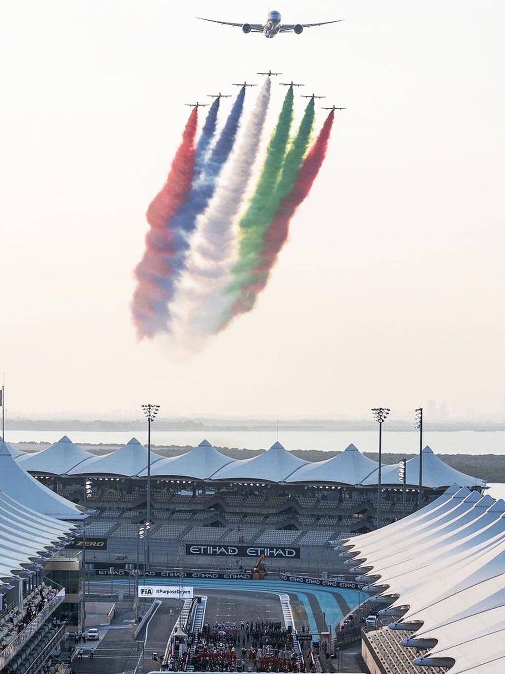 Colorful aircrafts flying over a race track at a motorsport event, creating smoke trails in red, blue, green, and white.