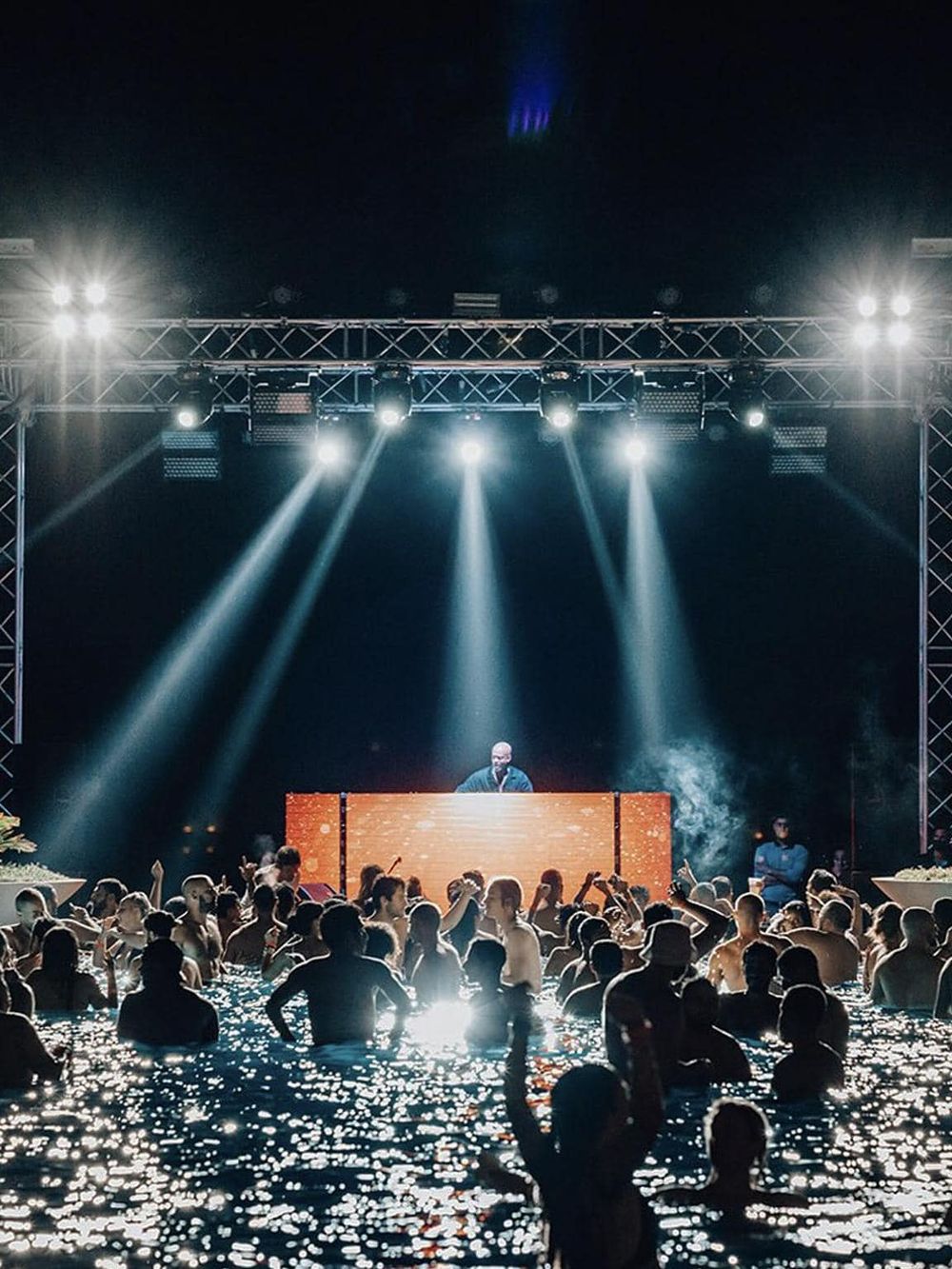 Vibrant night concert with DJ performing on stage, lively crowd in a water setting, bright stage lights, modern event atmosphere, QuestForDirections.