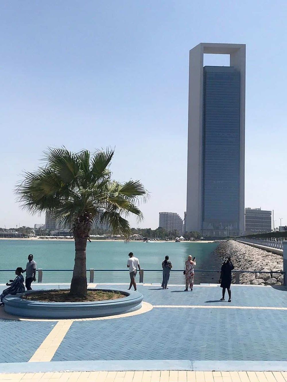 Modern skyscraper near waterfront with people enjoying outdoor scenery and palm trees, in Dubai.