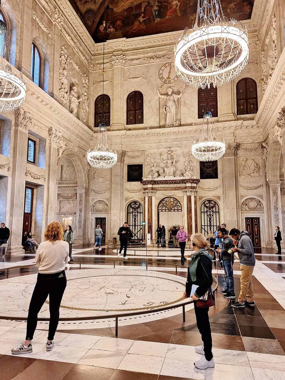 Intricate marble interiors of a grand historic building with chandeliers, featuring visitors exploring the majestic space.