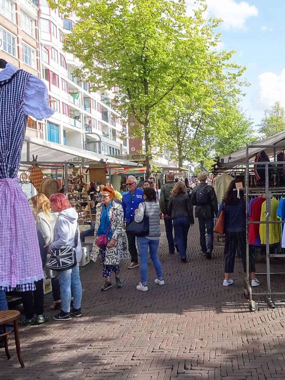 Vibrant outdoor street market with shoppers, colorful stalls, and green trees in a city setting.