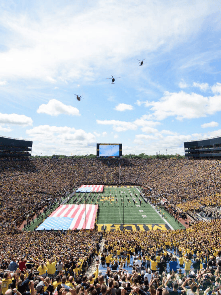 Crowd enjoying college football game at Michigan Stadium with national flags and helicopters overhead.