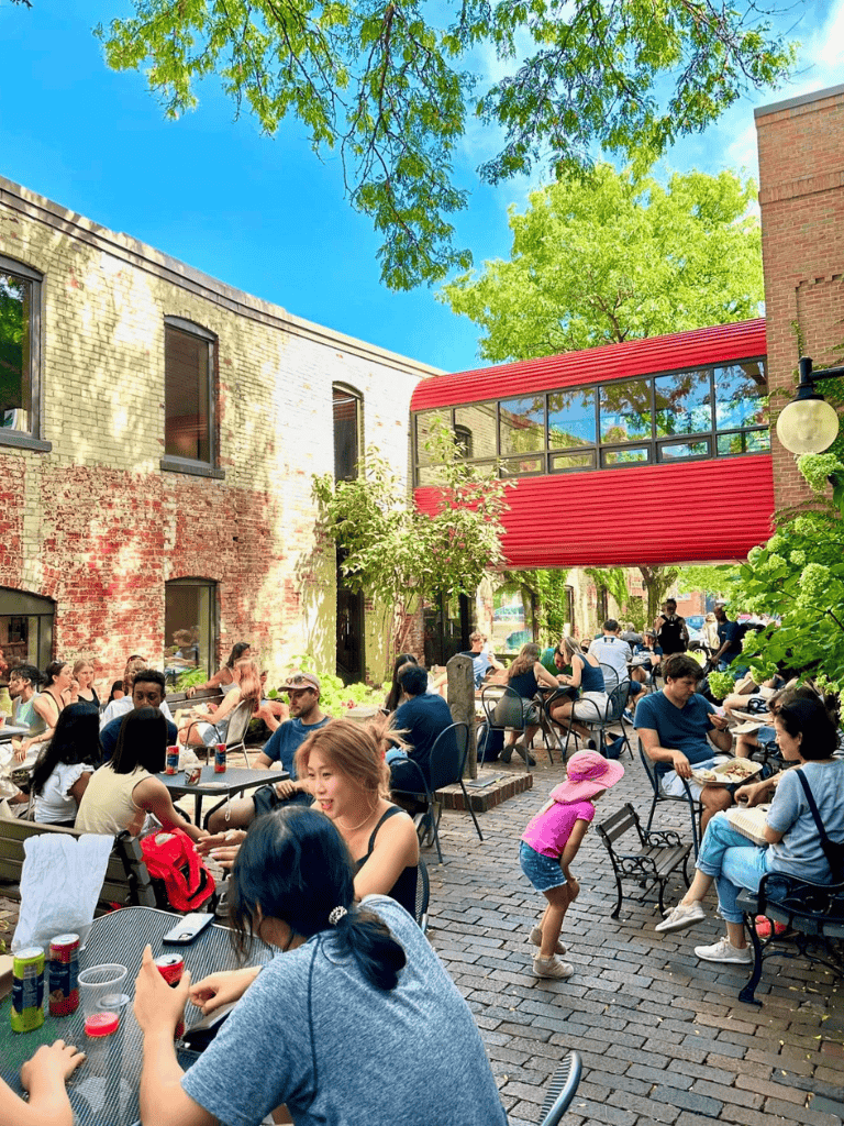 Outdoor urban cafe scene with people dining and children playing under a lush green canopy, vibrant and lively atmosphere.