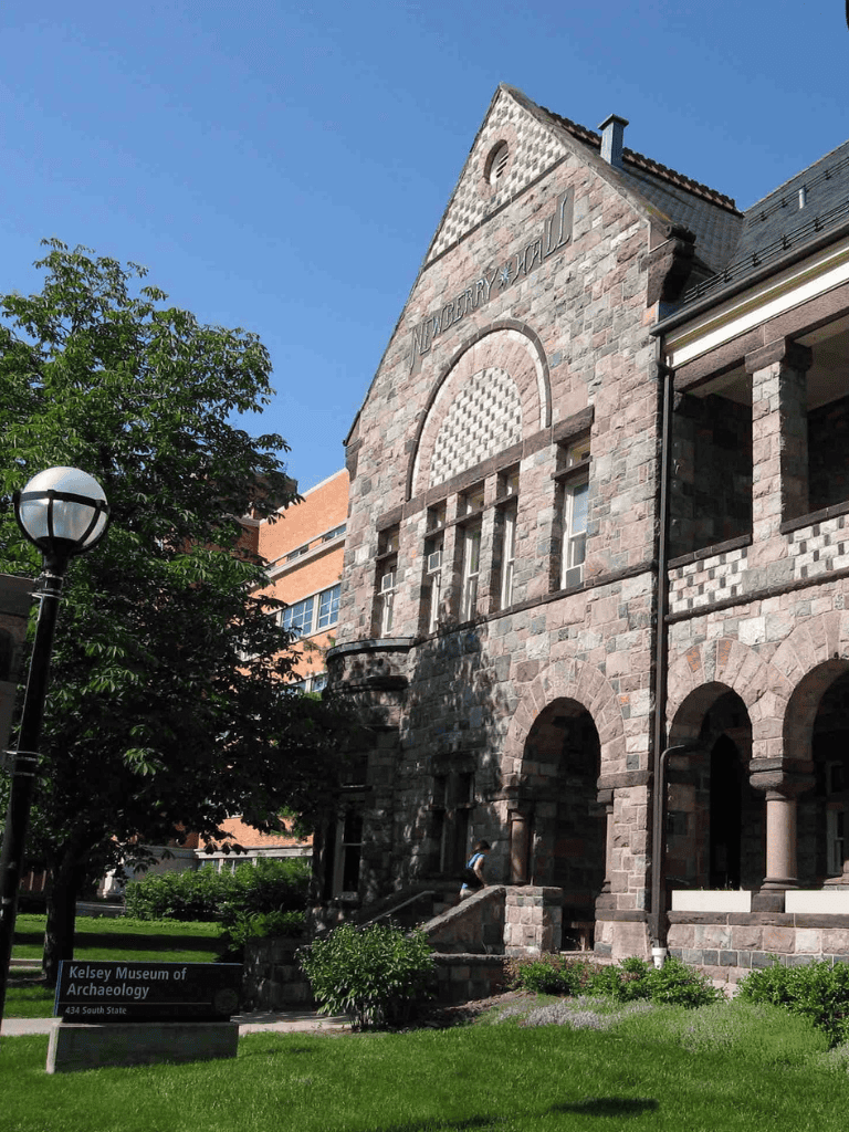 Stone building with arched windows and "Kelsey Museum of Archaeology" sign, sunny day.