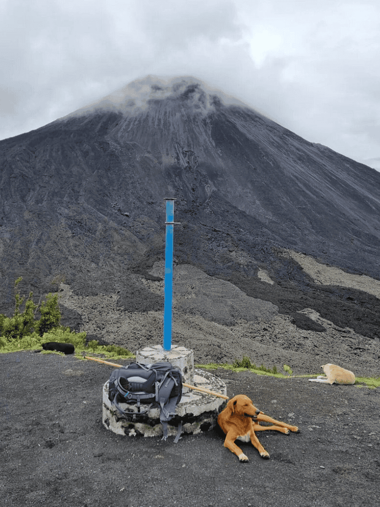 Volcano trekking scene with dogs resting near trail marker, cloudy mountain background, adventure travel, and outdoor exploration.
