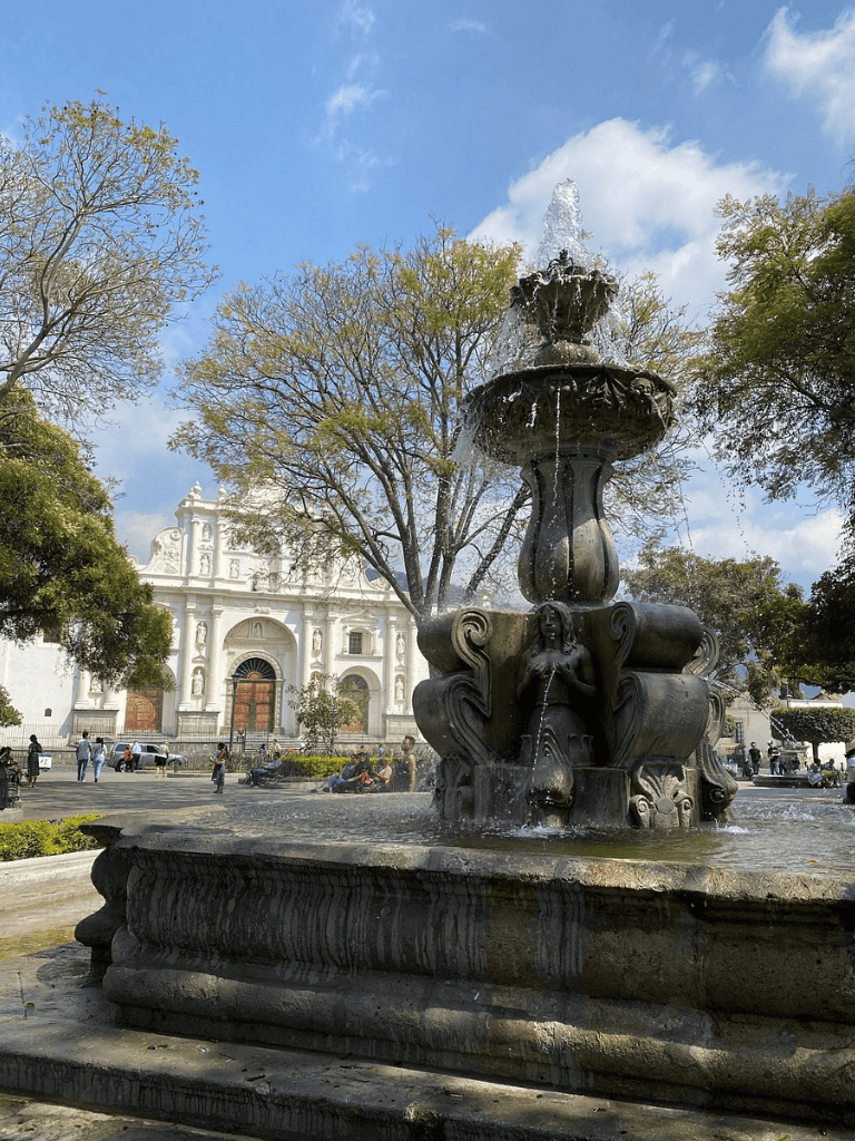 Historic fountain in a vibrant park, featuring intricate sculptures and surrounded by lush trees and a classic white church in the background.