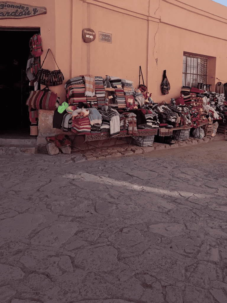 Colorful woven blankets and bags displayed outside a local shop in a rustic village setting.