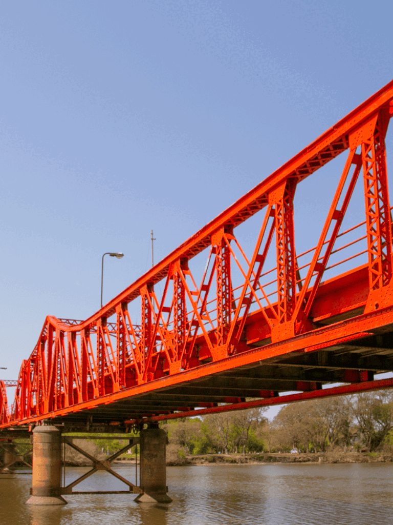 Red bridge over river with clear blue sky, scenic view, and lush greenery in the background.