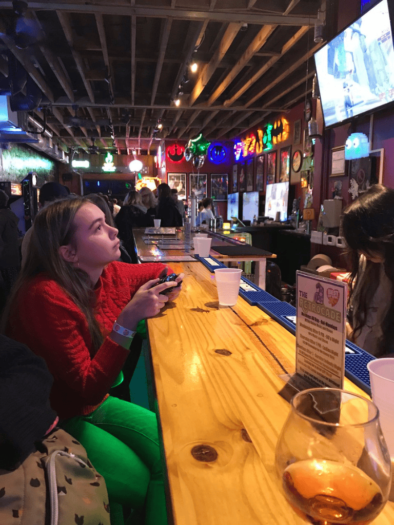 Colorful neon lights in a bar with patrons watching screens and a young girl in red and green seated at the bar.