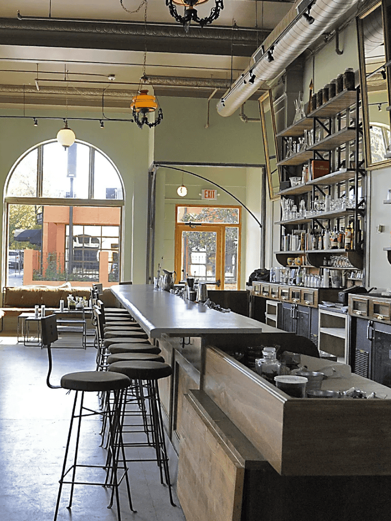 Cozy café interior with bar seating and large windows, stylish decor and bar shelves with bottles and glassware.