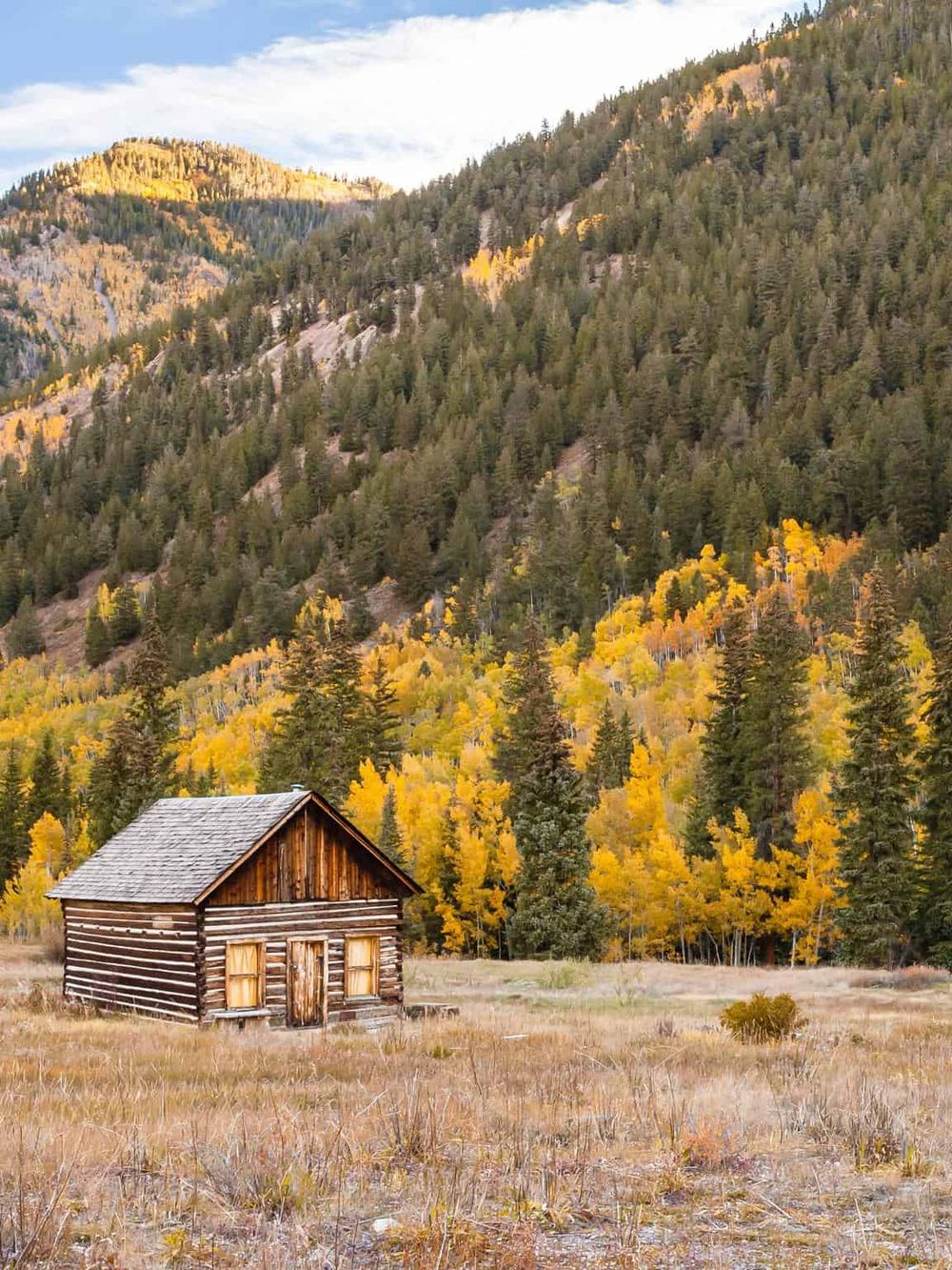 Rustic wooden cabin in a scenic mountain landscape with colorful fall foliage and evergreen trees.