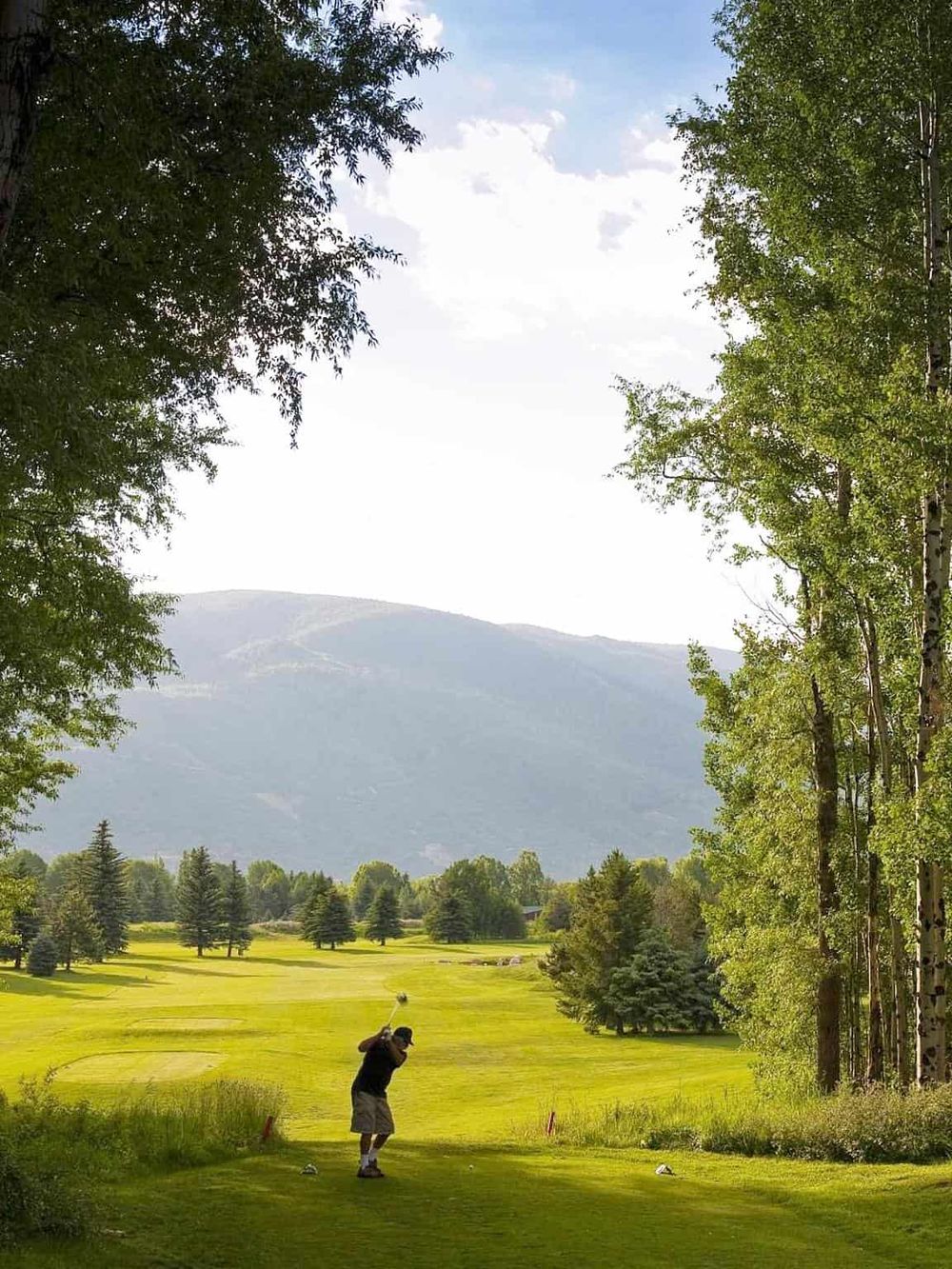 A scenic golf course with lush greenery, trees, and mountains in the background under a blue sky.