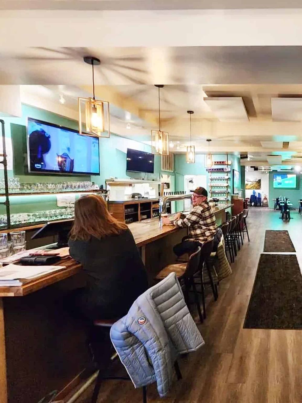 Cozy bar with TV screens, modern lighting, wooden counter, and patrons enjoying drinks in a stylish setting.