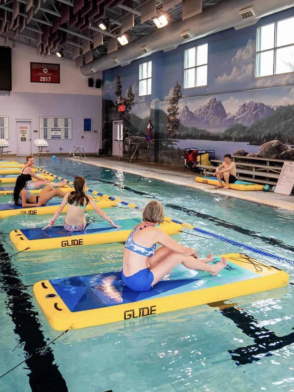 Indoor swimming pool with paddleboards, families enjoying water activities, scenic mountain mural background.
