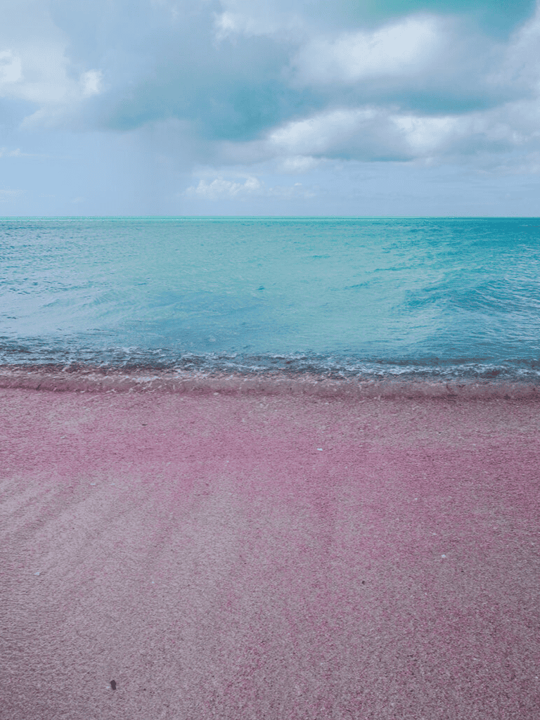 Calm turquoise ocean and pink sandy beach under cloudy sky.