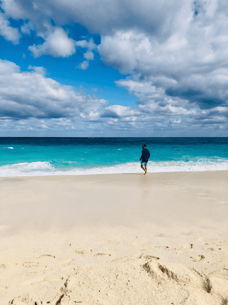 Serene beach scene with a person walking along the shore under cloudy sky, perfect for travel and vacation inspiration.