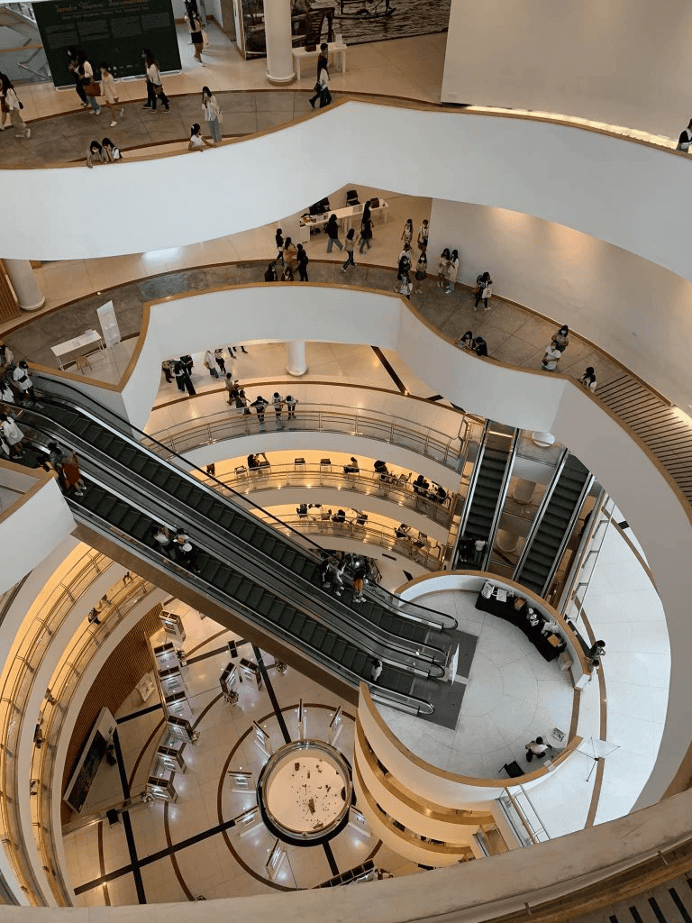 Indoor shopping mall with multiple floors, escalators, and shoppers in a modern, well-lit environment.