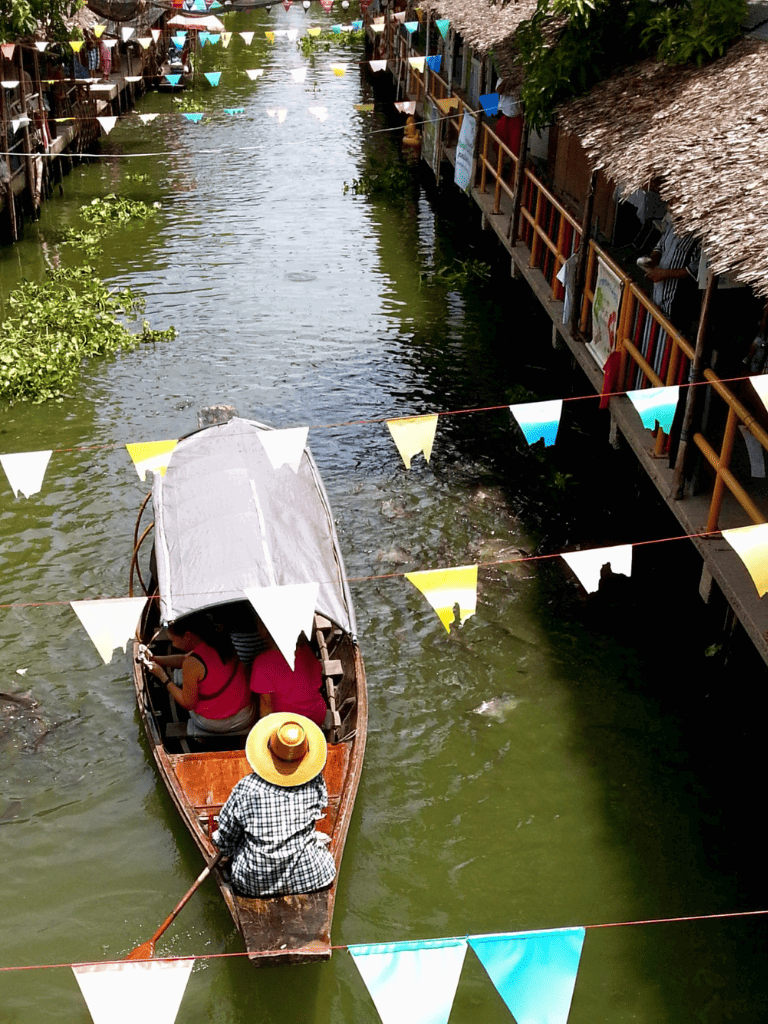Colorful boat navigating a narrow waterway between stilted cafes and shops, vibrant flags overhead, lively and scenic waterfront scene.