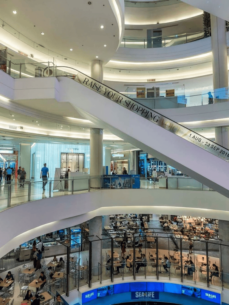 Bright multi-level shopping mall interior with escalators, shops, and dining area, modern design.