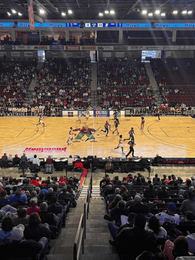 Basketball game at an indoor stadium, lively crowd, players in action, scoreboard visible, QuestForDirections branding.