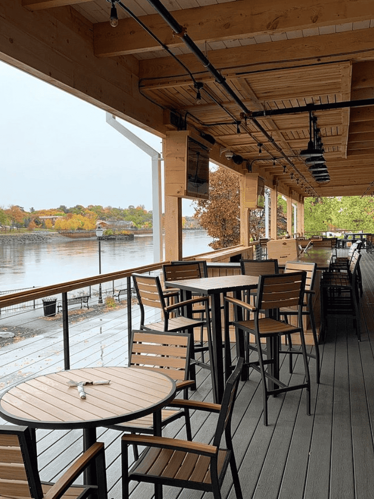 Outdoor waterfront dining area with wooden tables and chairs, overlooking a river with autumn foliage. Perfect for relaxing meals and scenic views.