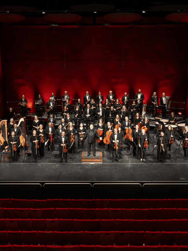 Orchestra performance on stage with conductor and musicians holding string, woodwind, brass, and percussion instruments.