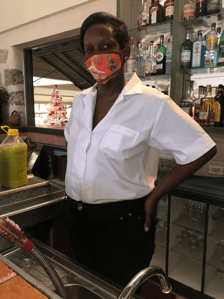 Bright woman in a white shirt and festive face mask behind bar with liquor bottles; holiday decor in background.
