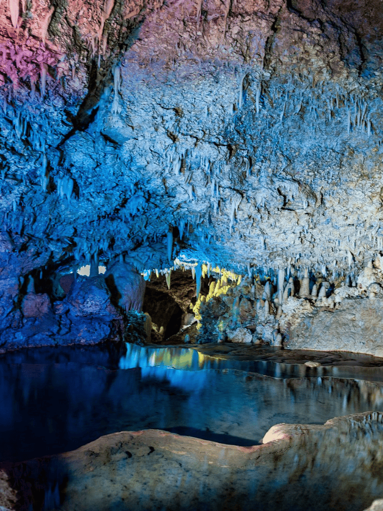 Colorfully illuminated cave with stalactites and stalagmites reflecting in underground water.