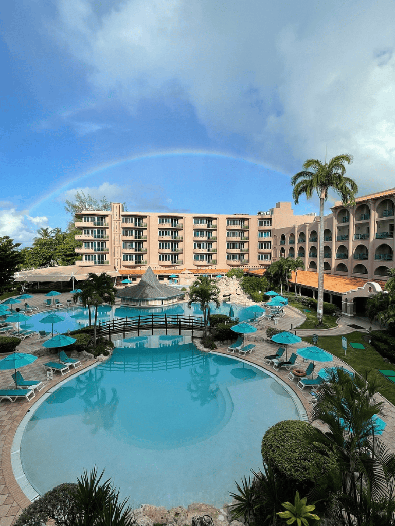 Colorful rainbow over tropical resort pool area, relaxing vacation destination, palm trees, blue umbrellas.