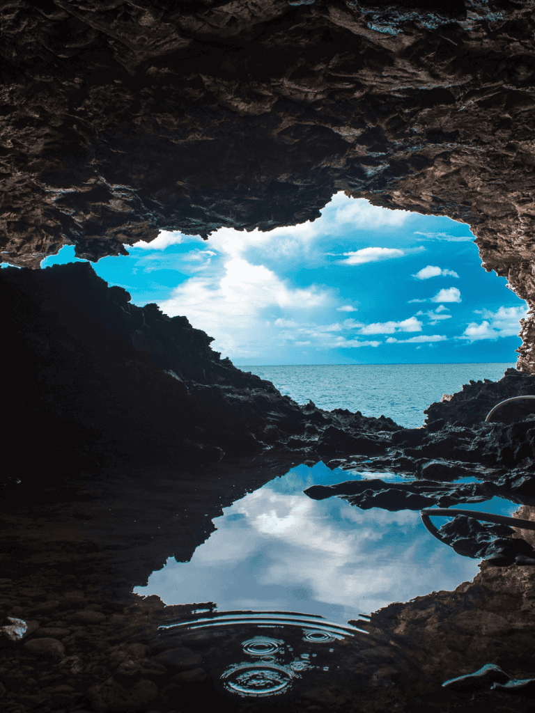 Seaside cave opening with view of ocean and blue sky, natural rugged coastline scenery.