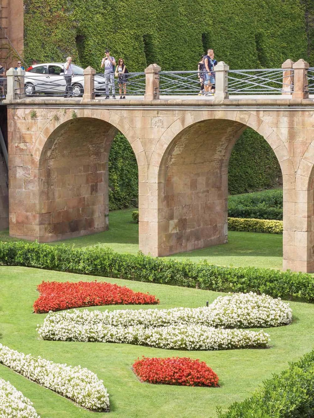 Bridge with pedestrians and parked cars in scenic park setting, surrounded by colorful flower beds and lush greenery.