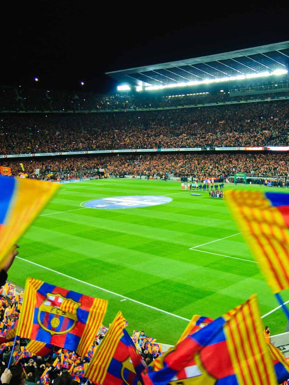 Vibrant football stadium filled with cheering fans, flags, and bright lights during a夜 game.