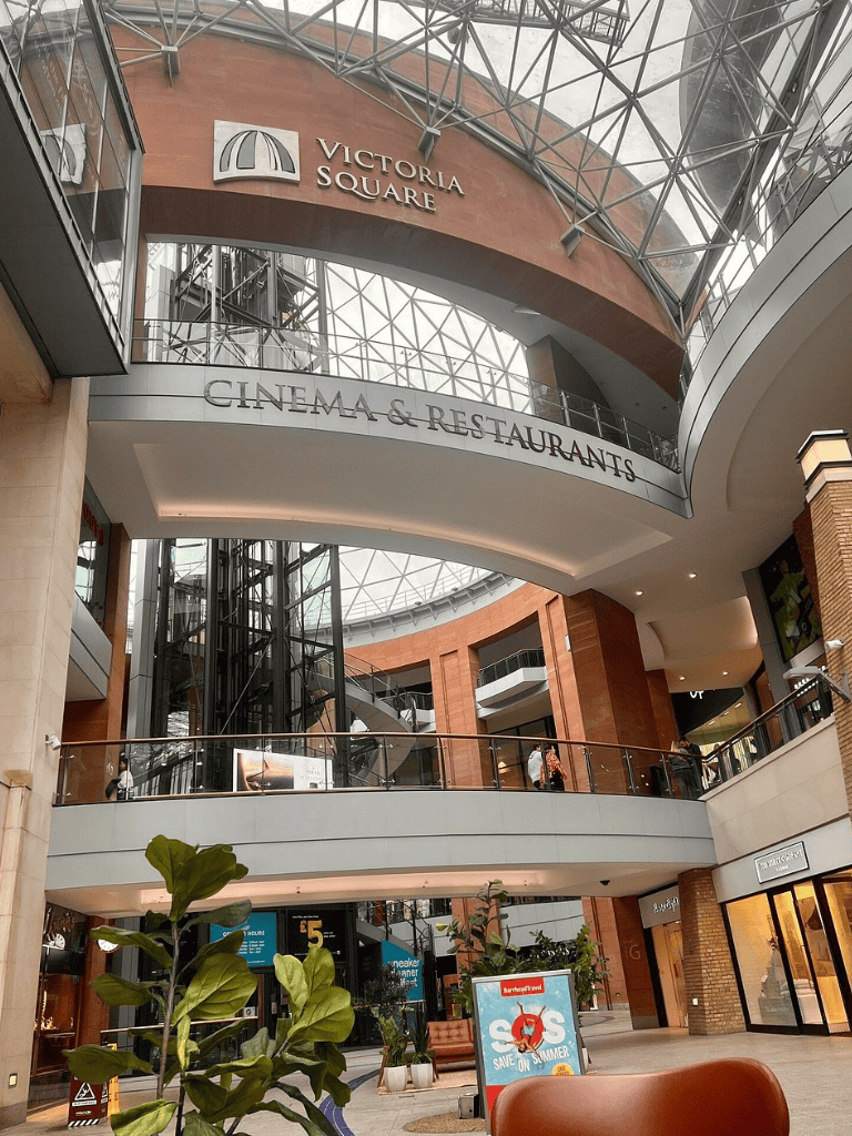 Bright indoor shopping mall with Victoria Square sign, cinema and restaurant signage, modern architecture, and greenery.