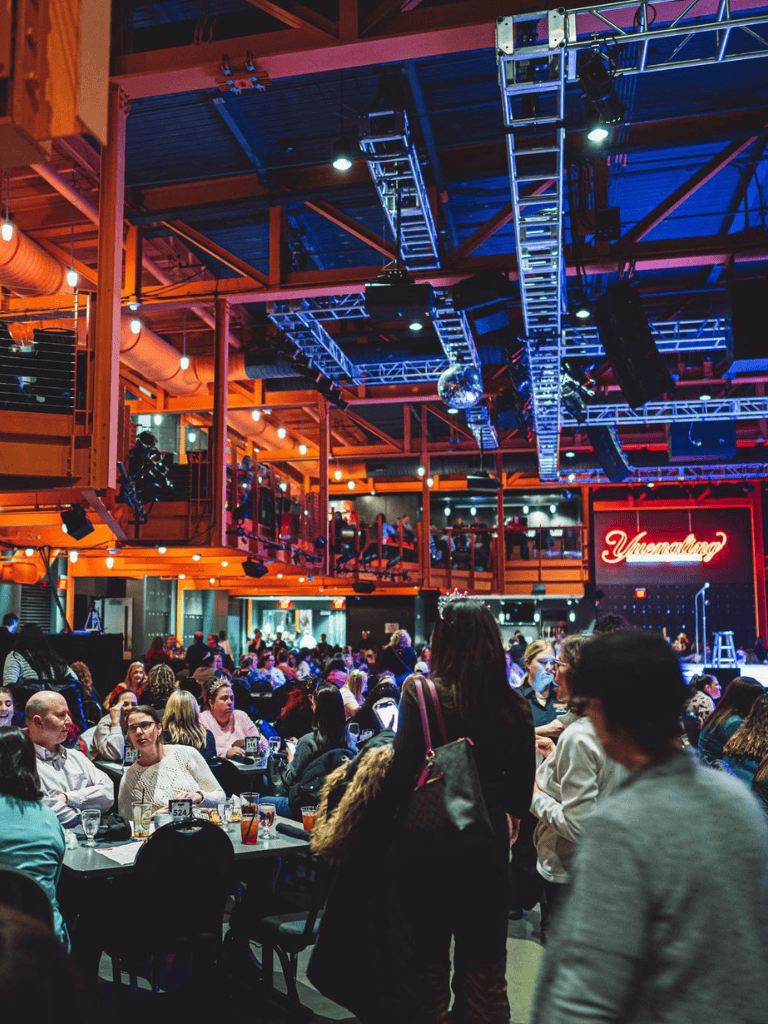 Brightly lit event venue with a lively audience, stage with neon sign, and colorful lighting effects.