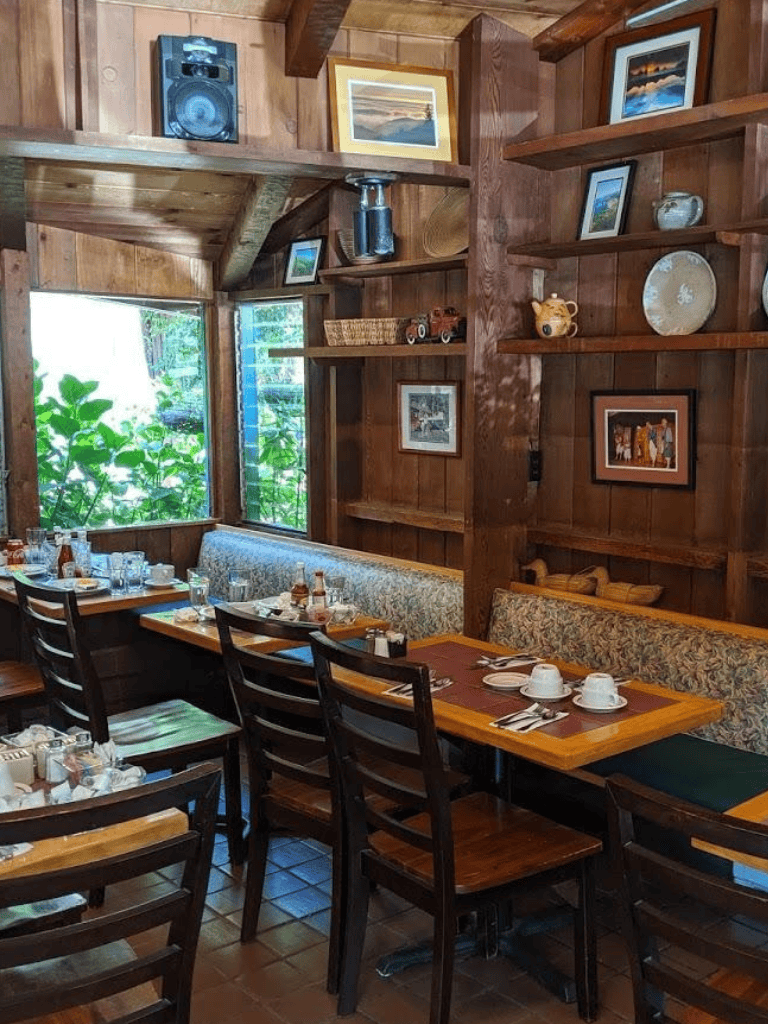 Cozy wooden restaurant interior with framed artwork and set table for dining, lush greenery visible outside the window.