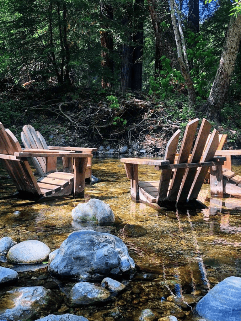 Adirondack-style wooden chairs in a clear mountain stream with rocks and lush green forest backdrop.