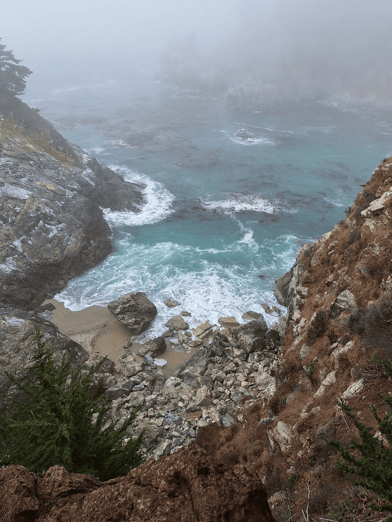 Serene rocky coast with ocean waves crashing between cliffs, foggy atmosphere, and coastal greenery.