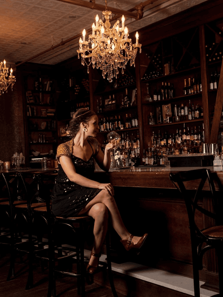 Elegant woman enjoying a cocktail at a sophisticated bar with crystal chandeliers and wooden shelves.